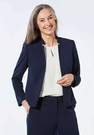 Blazer marine à coupe droite, à l'avant ouvert, avec des manches longues, associé à un haut blanc avec un détail en trou de clé et un pantalon assorti.