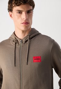 Sudadera con capucha en una mezcla de algodón verde olivo. Presenta una cremallera frontal, cordones y una etiqueta rectangular roja con el texto "HUGO".