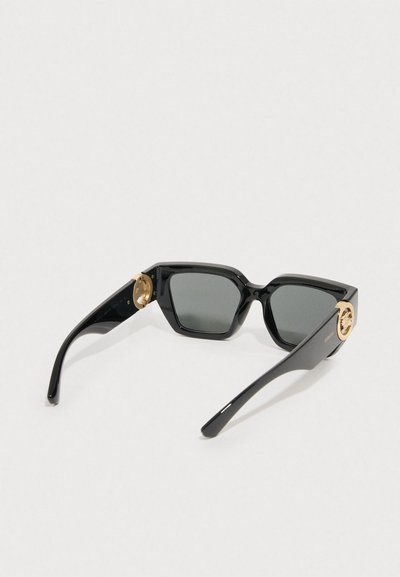 Mustade läätsedega musta ruuduga raamiga päikeseprillid, mille templettidel on kuldne Medusa embleem, lahtiselt valgel pinnal.