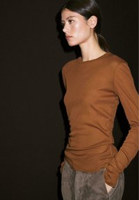 Woman with dark hair in a low ponytail wears a fitted brown long-sleeve shirt and gray pants, standing in profile against a dark background.