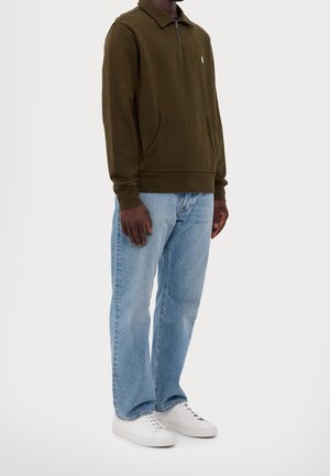 Homme portant un pull à fermeture éclair quart de couleur vert olive, un jean bleu clair et des baskets blanches, debout devant un fond clair uni.