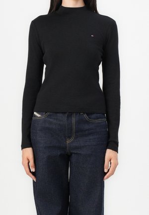 Vrouw draagt een effen zwart shirt met lange mouwen met een klein logo op de borst en donkerblauwe hoog taille jeans, tegen een witte achtergrond.