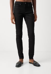Persona vestida con jeans ajustados negros y mocasines de cuero negros con detalle de hebilla dorada en forma de caballo, de pie contra un fondo blanco liso.