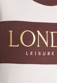 Camisa de manga corta blanca y burdeos con texto bordado en dorado que dice "LONDON LEISURE" en el pecho, mostrando una suave textura de tela.