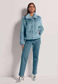 Helles, flauschiges, blaues Jackett mit Kragen, kombiniert mit türkisfarbenen Cordhosen und silbernen, spitzen Ankle-Boots, präsentiert an einem Modell vor einem neutralen Hintergrund.