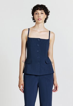 Donna con capelli corti e ricci, che indossa un top senza maniche blu navy con bottoni e pantaloni abbinati, in piedi davanti a uno sfondo bianco semplice.