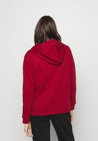Felpa rossa realizzata in tessuto morbido, con tasca a marsupio e cappuccio regolabile con coulisse. Il modello indossa pantaloni neri.