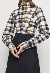 Plaid shirt with a high collar in navy, white, and yellow tones. Features long sleeves, button details, and a fitted design.