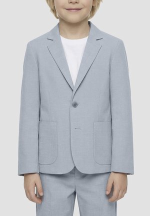 Malo dijete obučeno u svijetloplavi sako i odgovarajuće hlače preko bijele košulje, stoji ispred jednobojne svijetle pozadine.