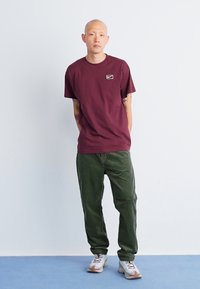 Burgundy short-sleeve t-shirt with a small Nike logo, paired with textured green corduroy pants and light sneakers on a blue surface.