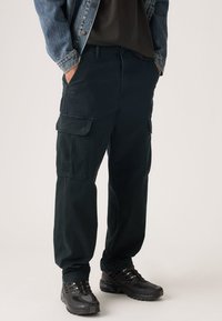 Μαύροι cargo παντελόνια με πλαϊνές τσέπες, κατασκευασμένα από μαλακό ύφασμα, εφαρμοστή γραμμή, σε συνδυασμό με υφασμένα μαύρα αθλητικά παπούτσια και τζιν μπουφάν.