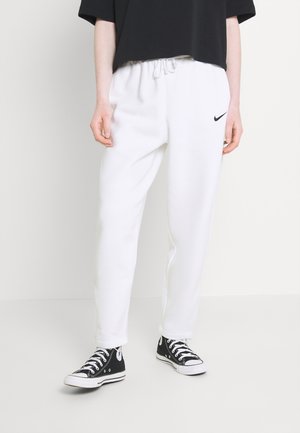 Persoon die witte Nike sweatpants en zwarte hoge sneakers draagt, staat tegen een effen witte achtergrond.