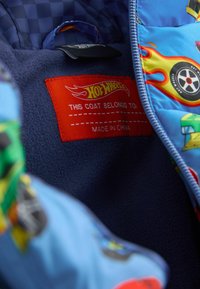 Interior de un abrigo acolchado azul con gráficos de coches coloridos, una etiqueta roja que dice "Hot Wheels" y un forro suave de polar marino.