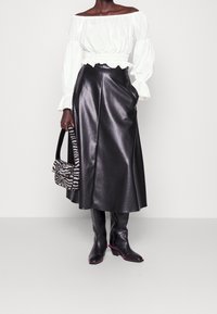 Blusa bianca off-the-shoulder con maniche a sbuffo, abbinata a una gonna midi in pelle ecologica nera. Outfit accessoriato con una borsa a fantasia bianco e nero.