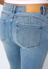 Ljusblå denimjeans med en slät textur. Har en läderlapp med texten "NOISY MAY" och två bakfickor med sydda kanter.