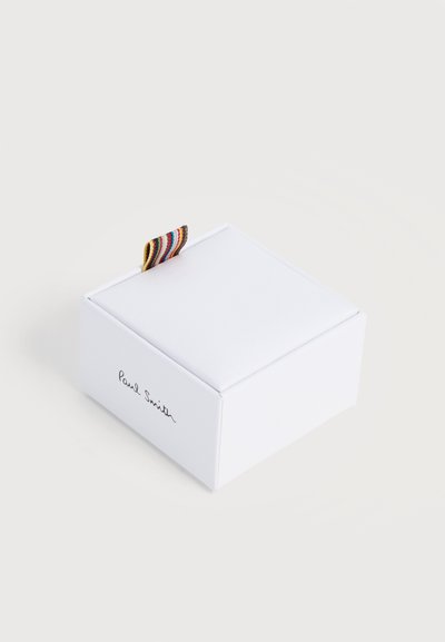 Boîte carrée blanche Paul Smith avec une languette en tissu rayé multicolore sur un fond clair uni.