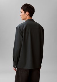 Camicia verde scuro con vestibilità comoda, maniche lunghe e polsini con bottoni, realizzata in tessuto liscio. Presenta un colletto classico e un pezzo dietro. 