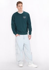 Felpa color teal con colletto a girocollo e piccola grafica bianca sul petto, abbinata a pantaloni cargo azzurri chiari con molteplici tasche.