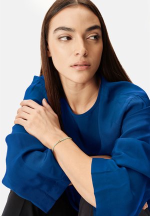 Blouse bleu roi avec des manches amples et volantées. Un bracelet délicat avec des perles vertes et un accent doré au poignet. Fond neutre.