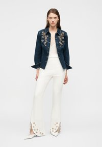 Γυναίκα που στέκεται φορώντας ένα σκούρο τζιν μπουφάν με διακοσμητικά από χάντρες σε σχέδιο λουλουδιών, λευκή δαντέλα στην κορυφή και λευκό παντελόνι με φαρδιά πόδια με λουλουδένιες λεπτομέρειες στα τελειώματα.