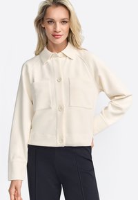 Crème-kleurige button-up shirt met kraag, twee borstzakken en lange mouwen. Glad materiaal met een iets cropped pasvorm.