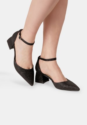 Zapatos de tacón negros con purpurina, punta afilada y un tacón bloque bajo. Cuentan con una correa ajustable en el tobillo. La textura es brillante y suave.