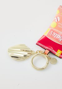 Gold metal keychain with a textured, wavy disc and a circular ring; packaging in vibrant red and peach tones featuring colorful designs.