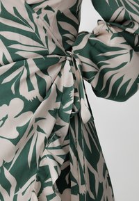 Zielono-kremowa tkanina w kwiatowy wzór z drapowanym fasonem, falbaniastymi brzegami i wiązanym detalem w pasie. Gładka tekstura i lekki materiał.