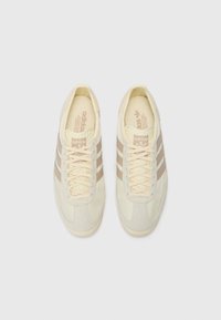 Sneakers w kolorze kremowym z trzema beżowymi paskami, uszyte z teksturowanej zamszowej cholewki, płaskimi sznurowadłami i wyściółką podeszwy. Klasyczne logo Adidas na języku.