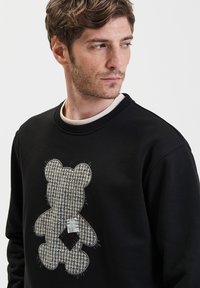 Zwart sweatshirt met een groot berenontwerp in een gestructureerde grijze stof met houndstooth patroon en geborduurde accenten.