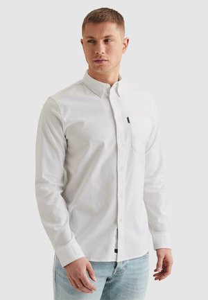 Moški, oblečen v oprijeto belo srajco z gumbi in prsnim žepom, oblečen v svetlo modre kavbojke, stoji pred enobarvno ozadjem.