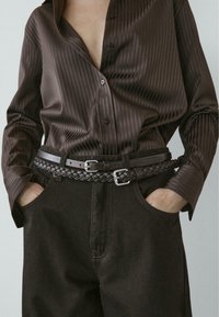 Camicia a righe marrone con scollo a V profondo, abbinata a pantaloni a gamba larga marrone scuro. La cintura nera intrecciata con fibbia argentata accentua il look.