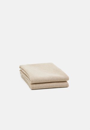 Couverture en laine beige pliée avec un motif chevrons subtil, posée sur un fond blanc.