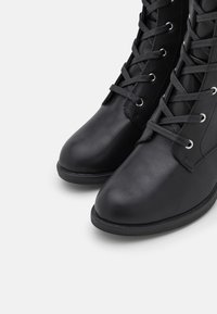 Czarne skórzane buty z zaokrąglonym noskiem, z płaskimi szarymi sznurowadłami, metalowymi oczkami i teksturowaną powierzchnią. Solidna czarna gumowa podeszwa.