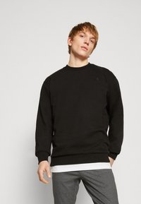 Felpa nera con scollatura a girocollo e polsini a costine, realizzata in tessuto morbido. Presenta un piccolo logo sul petto e una vestibilità comoda.