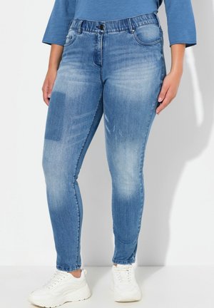 Jeans ajustados en denim de color azul claro con un ligero desgaste. Presentan un cierre de botón y bolsillos frontales. Combinados con zapatillas blancas.