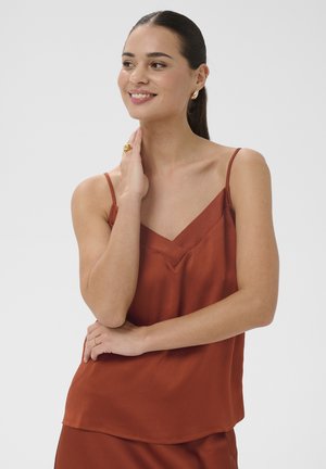 Femme souriante aux cheveux foncés portant un haut sans manches couleur rouille, touchant son cou d'une main et croisant les bras confortablement.