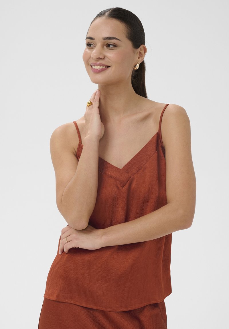 Femme souriante aux cheveux foncés portant un haut sans manches couleur rouille, touchant son cou d'une main et croisant les bras confortablement.