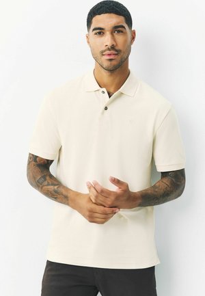 Beige polo shirt with a classic collar, two-button placket, and a subtle logo on the chest; smooth texture and short sleeves.