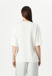 Cream, short-sleeve blouse with a straight cut, soft texture, and center back seam, paired with light gray pants. Minimalist design.