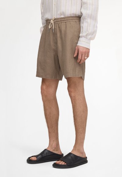 Homme portant un short beige à cordon de serrage, une chemise à manches longues rayée et des sandales noires à bout ouvert, debout devant un fond blanc.
