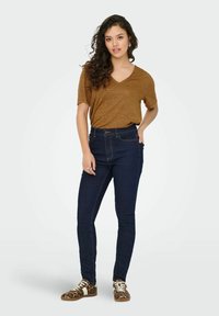 Donkerblauwe high-waisted jeans gecombineerd met een bruine V-hals T-shirt en sneakers met dierenprint. Het model heeft losjes krullend haar en een neutrale achtergrond.