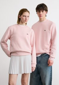 Νεαρός άνδρας και γυναίκα στέκονται, και οι δύο φορούν ανοικτό ροζ φούτερ με μικρά πράσινα λογότυπα· η γυναίκα φοράει λευκή πλισέ φούστα, ο άνδρας μπλε τζιν.