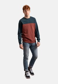 De hoodie heeft een marineblauw bovenstuk, een roestkleurig onderstuk, lange mouwen, ribgebreide manchetten en een ronde halslijn. Draagt met lichte jeans en hoge sneakers.