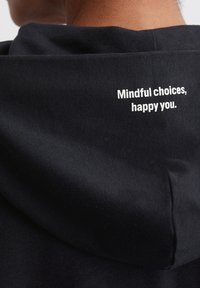 Zwarte hoodie van zachte stof, met een relaxte pasvorm en een bedrukt bericht op de achterkant: "Bewuste keuzes, gelukkig jij."