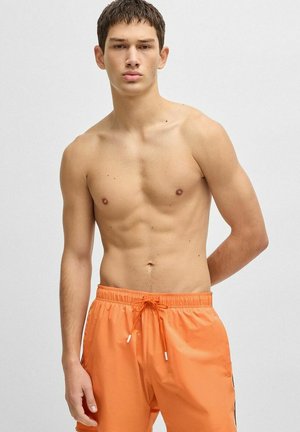 Junger Mann mit kurzen dunklen Haaren, der vor einem schlichten Hintergrund steht, oberkörperfrei, trägt orangefarbene Badeshorts mit Kordelzug.