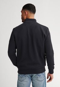 Felpa con zip di colore blu navy con un motivo a diamante testurizzato, polsini e orlo a coste, abbinata a jeans blu chiaro. Vista posteriore.