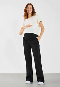 Umstandsmode bestehend aus einem locker sitzenden, kurzärmeligen cremefarbenen Oberteil und schwarzen Hose mit elastischem Bund und Taschen, kombiniert mit weißen Schuhen.