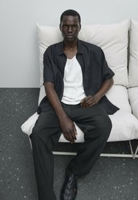Camicia nera a maniche corte sopra un canottiera bianca, abbinata a pantaloni neri larghi e sandali neri intrecciati, seduto su un divano di colore chiaro.