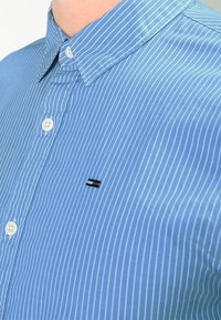 Světle modrá pruhovaná košile s límcem na knoflíky, zdobená malým logem na levé části hrudi v červené, bílé a černé barvě.
